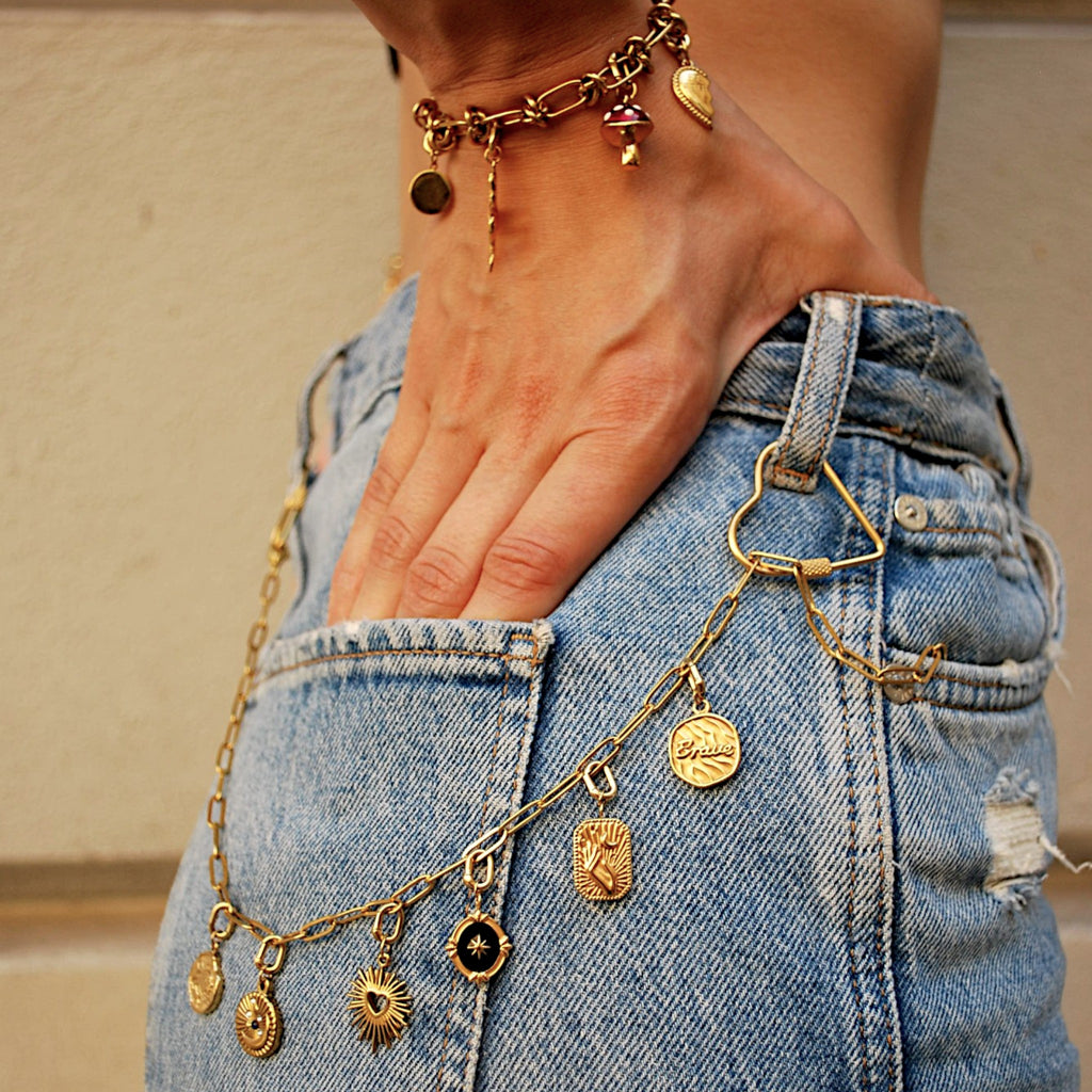 Złoty charms Sacred Heart marki HOOKS przypięty do łańcuszka przy jeansach, wykonany ze stali nierdzewnej.